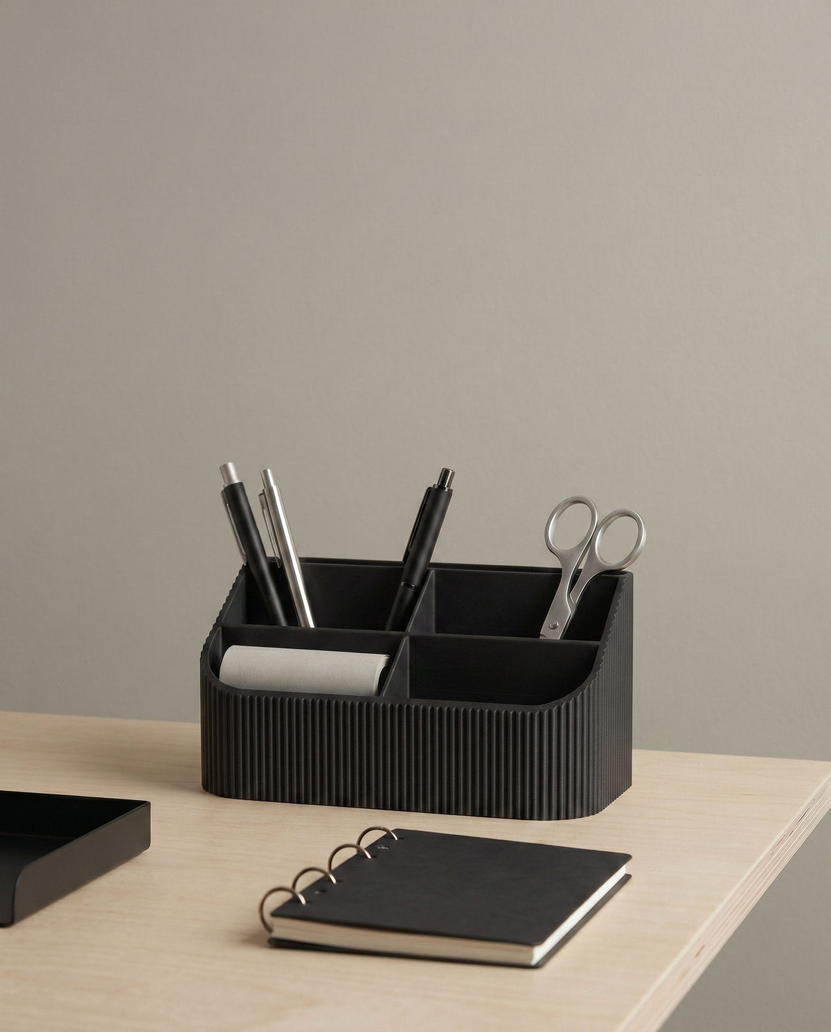 COCO Black Wood minimalist black wood desk organizer on a light ash wood table. The multi-compartment tidy holds pens and metal scissors, alongside a black spiral notebook and a matte black desk lamp, showcasing a sleek and organized workspace.