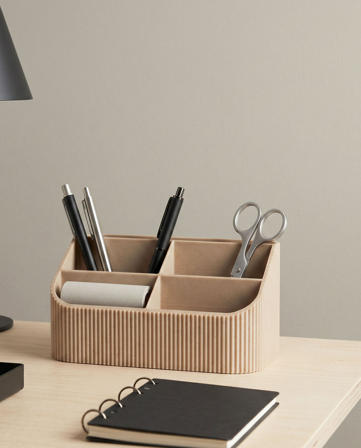 COCO minimalist wood desk organizer on a light ash wood table against a soft green wall. The multi-compartment tidy holds pens and metal scissors, alongside a black spiral notebook and a matte black desk lamp, showcasing a sleek and organized workspace.