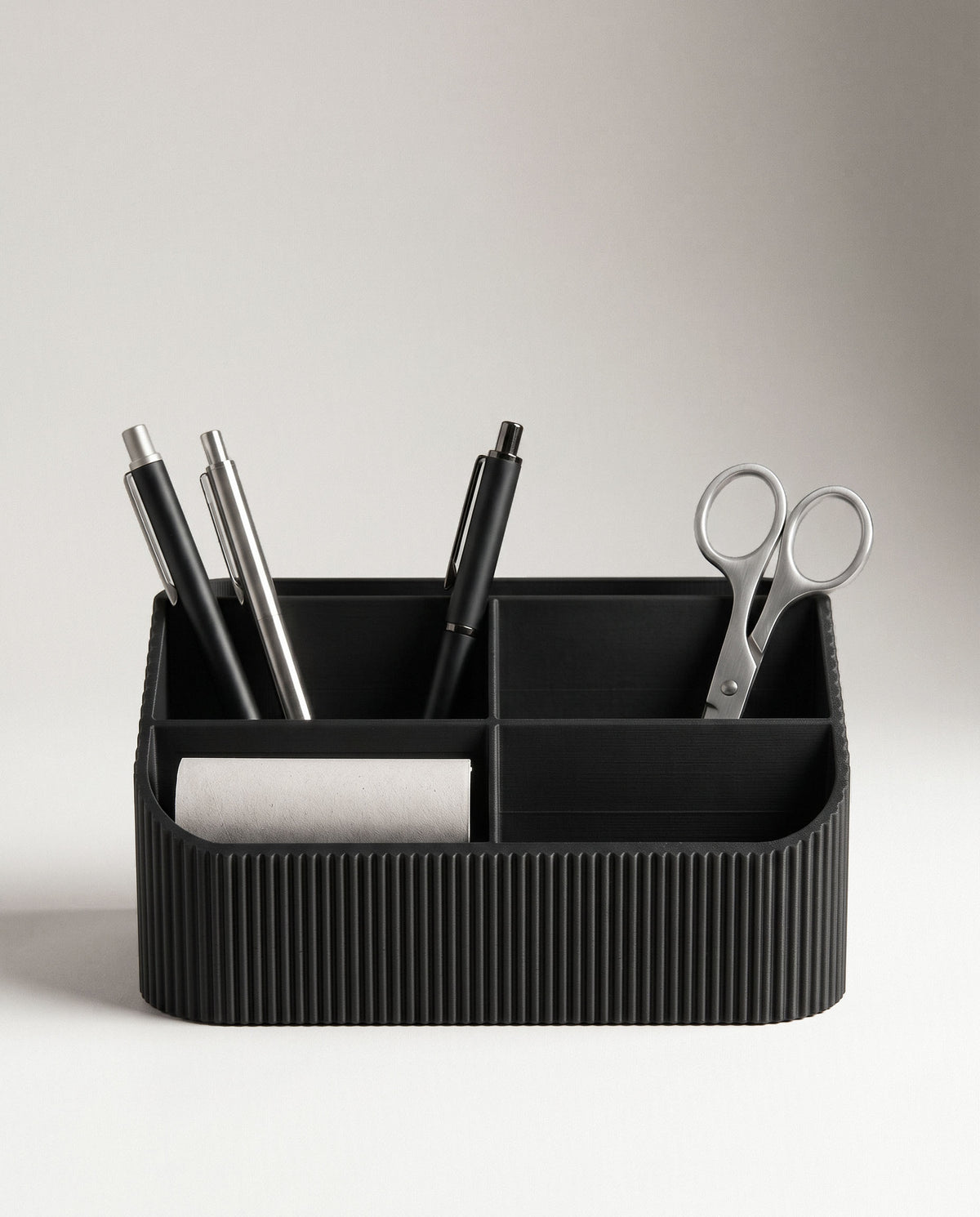 A clean, front-facing portrait of the COCO black fluted wood desk organizer against a seamless off-white background. The compartments are neatly filled with three pens and a pair of polished metal scissors, highlighting its ribbed texture.