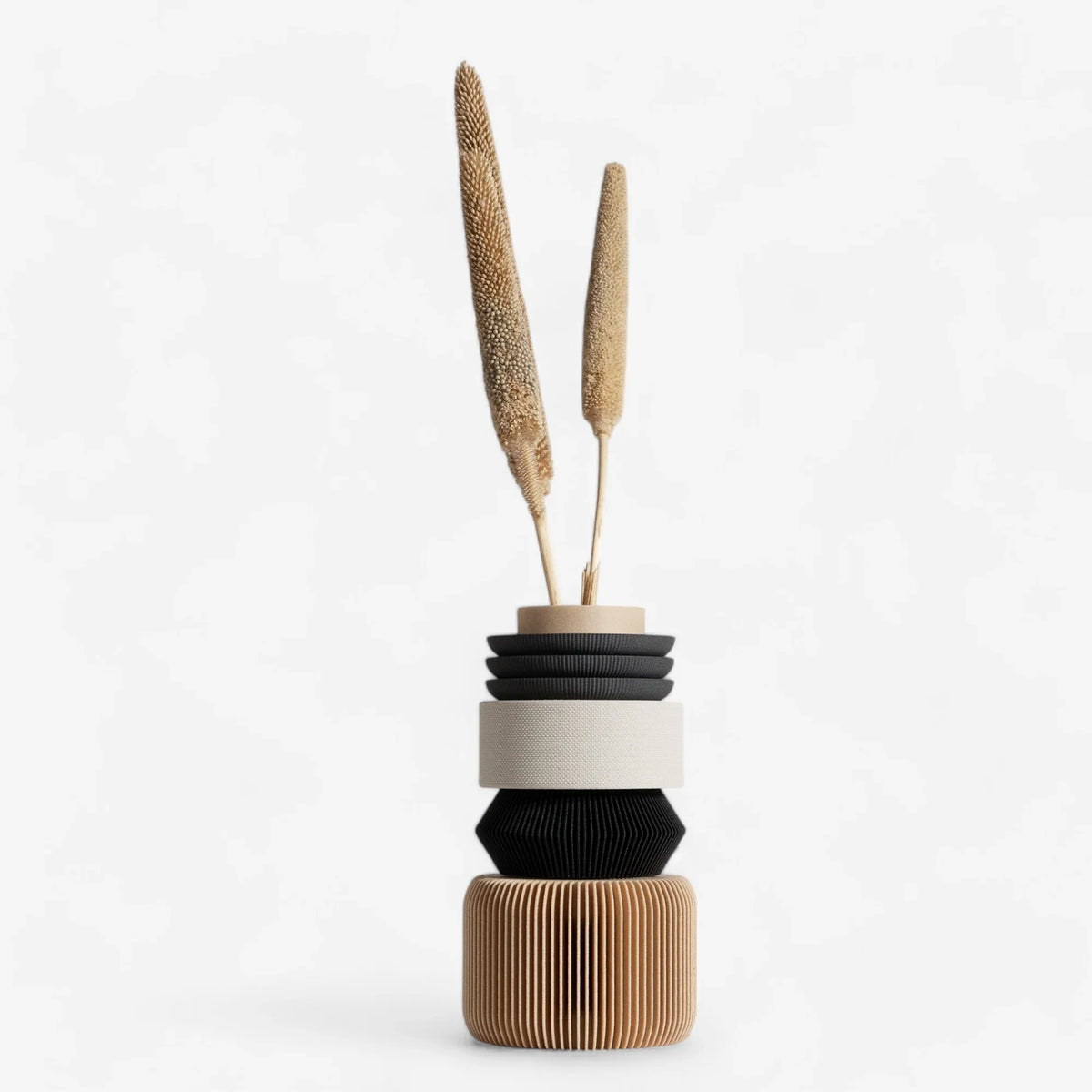 A modular vase made of multiple geometric shaped parts in black, white, beige, and brown colors, with two dried plants inserted on top.