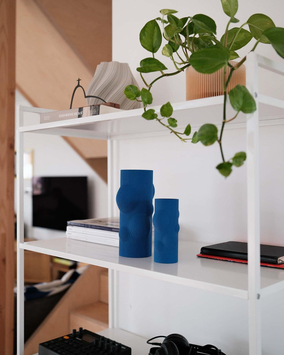 Blue cylindrical vase on white shelving unit with objects, books, and a plant in a room.