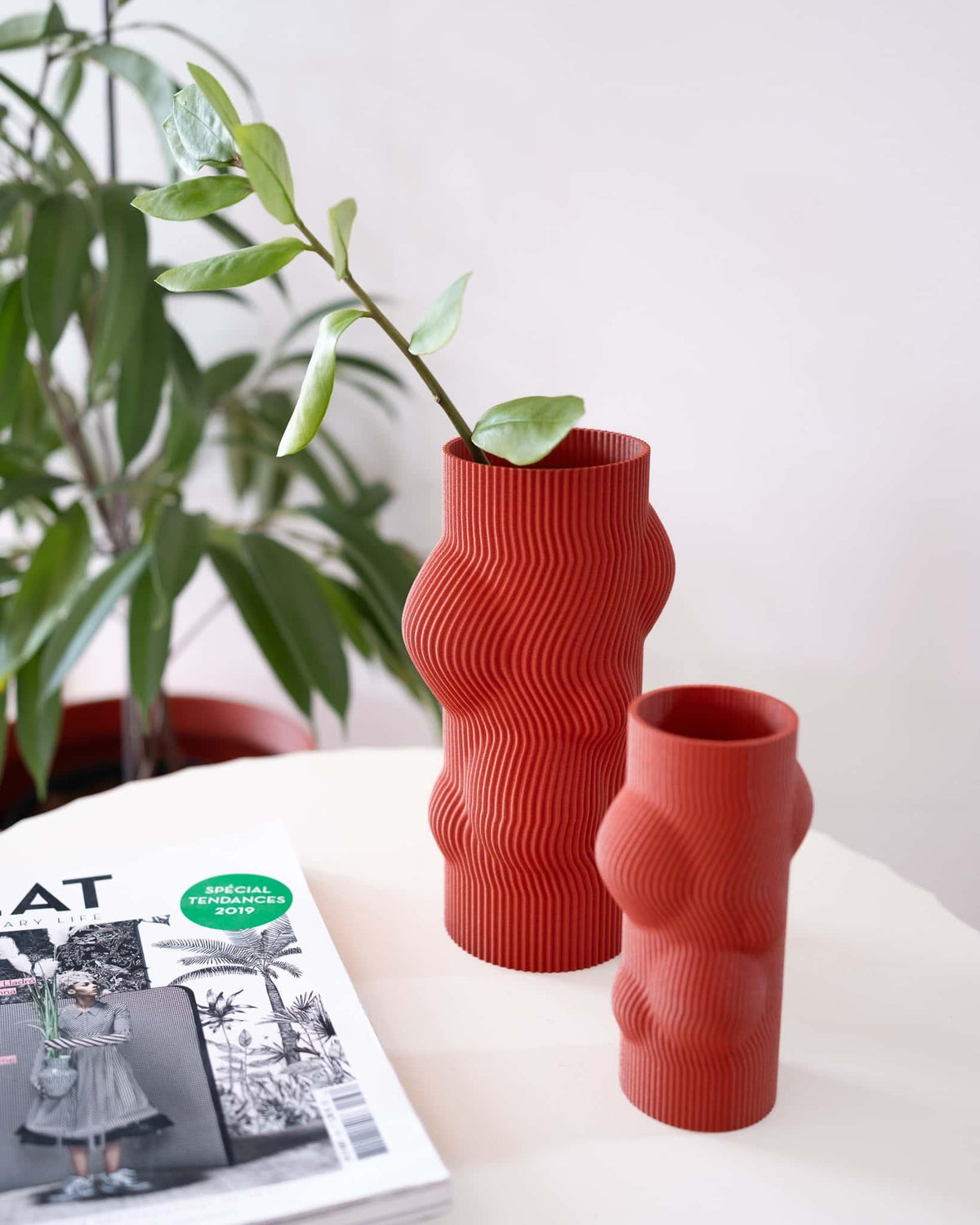 Two red textured vases on a table with a plant and magazine in the background.
