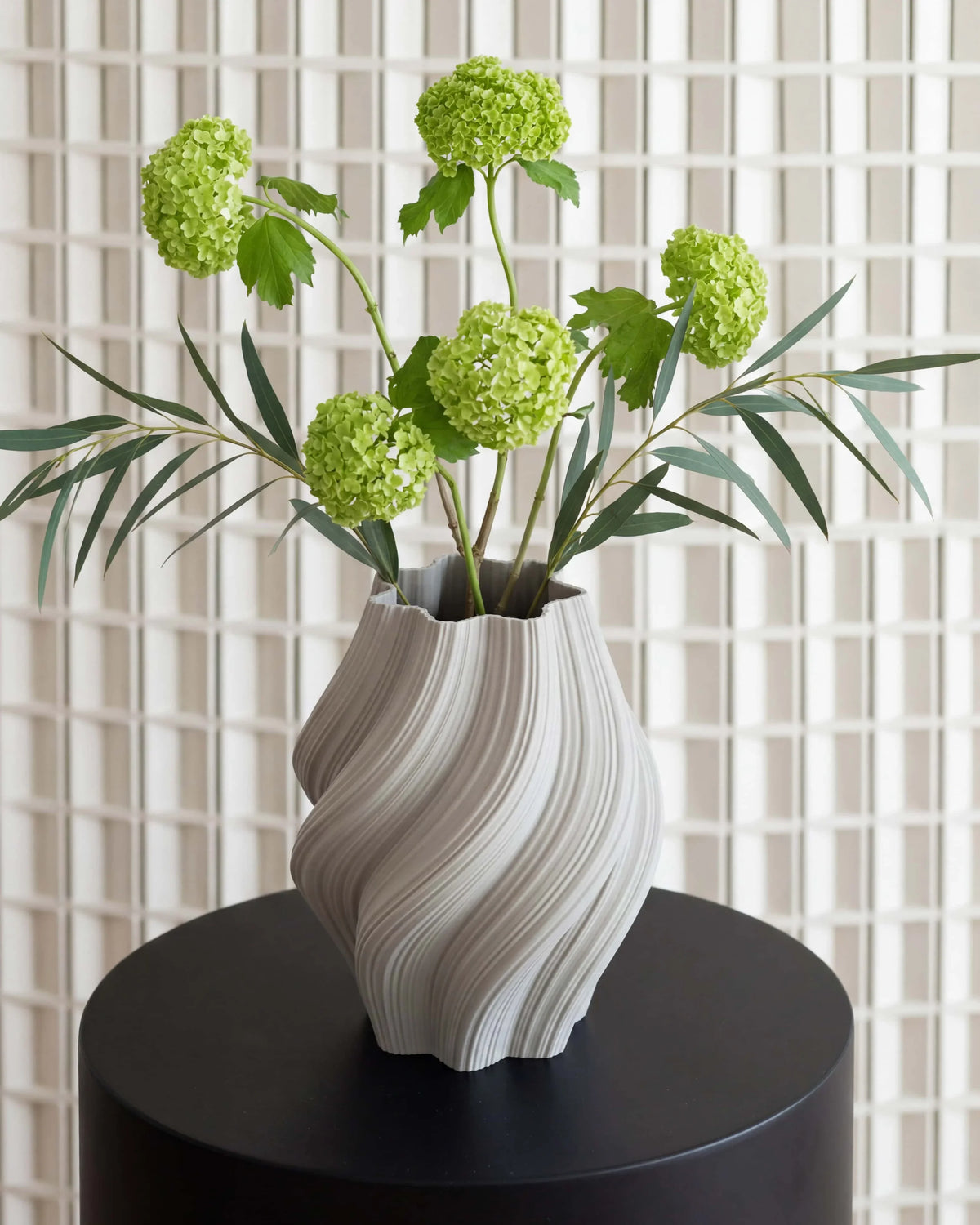 Crease white vase 22cm - Decorative vase with greenery on a black surface against a tiled wall.