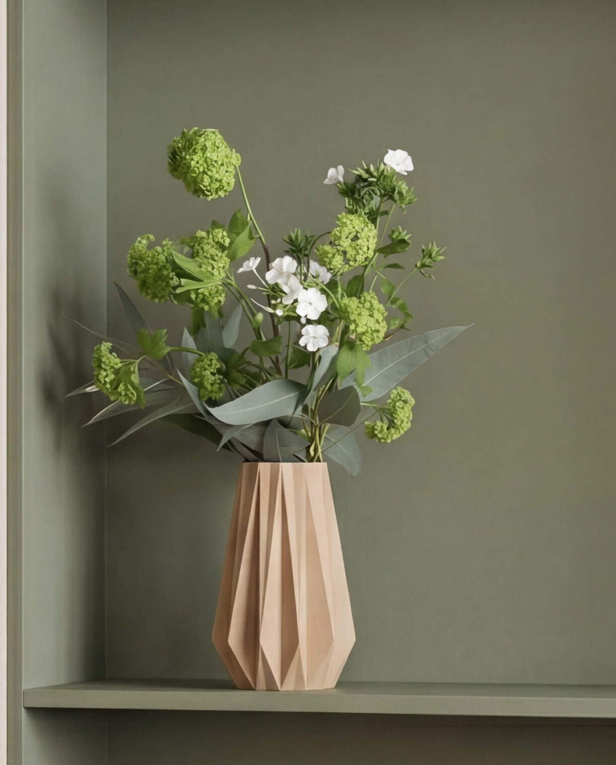 Origami vase natural color with green and white flowers on a green shelf against a green wall