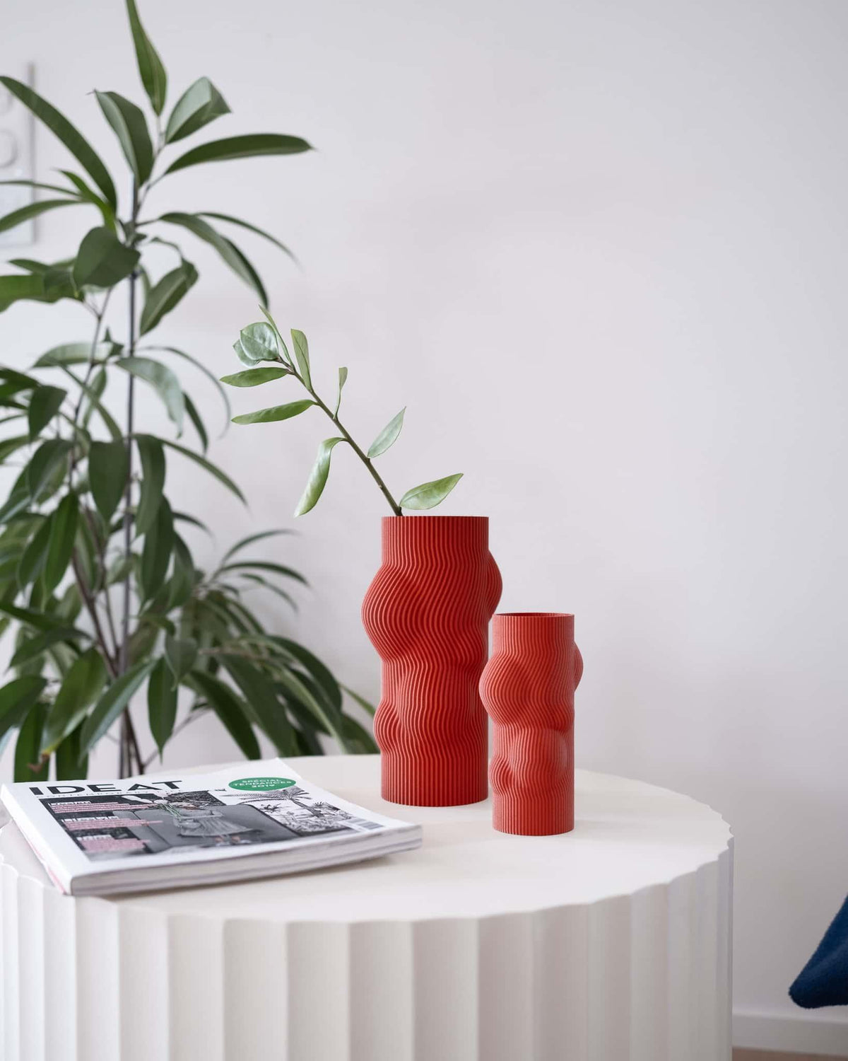 Two red textured vases on a white table with a plant and magazine in the background.