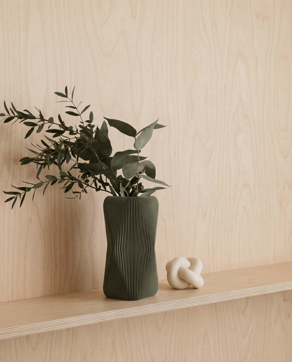 olive green vase with plants on a wooden shelf against a light wood panel wall